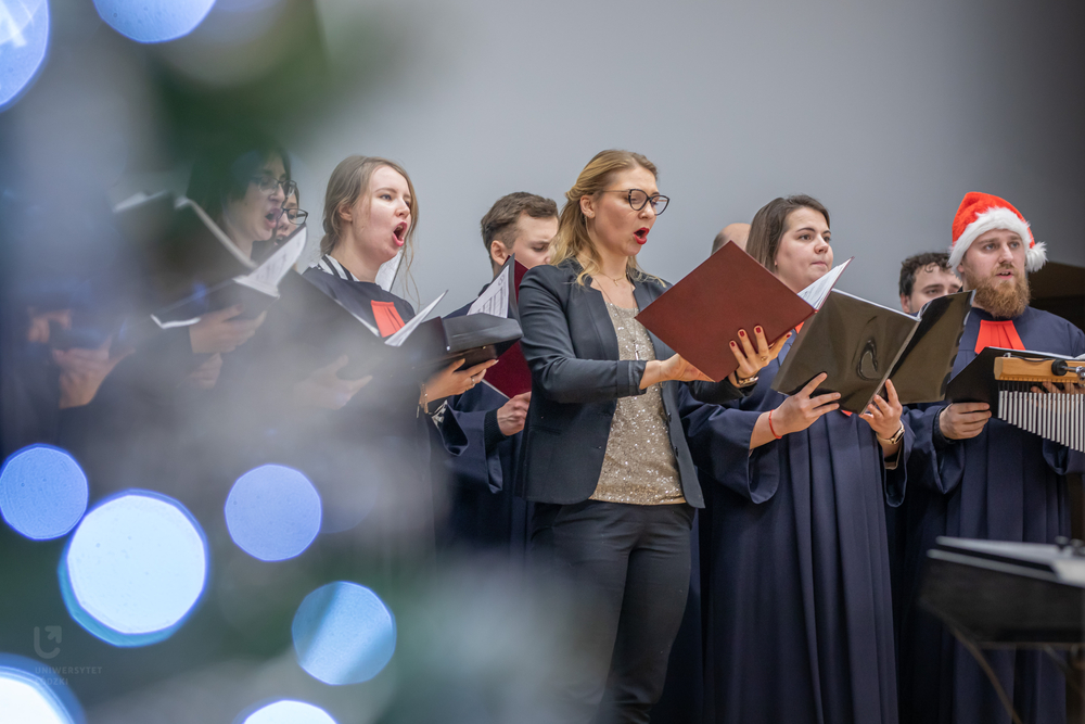 Members of the University of Lodz Choir singing during a concert. Blurred Christmas tree lights are visible