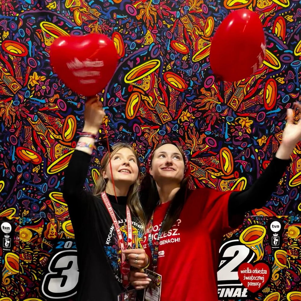 2 young women holding red heart-shaped balloons with the words "Great Orchestra of Christmas Charity" written on them